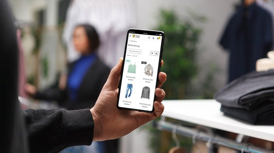 Omnichannel-Service: Im stationären Bekleidungsgeschäft hat ein Kunde die Mode-Artikel im Onlineshop auf seinem Smartphone in der Hand