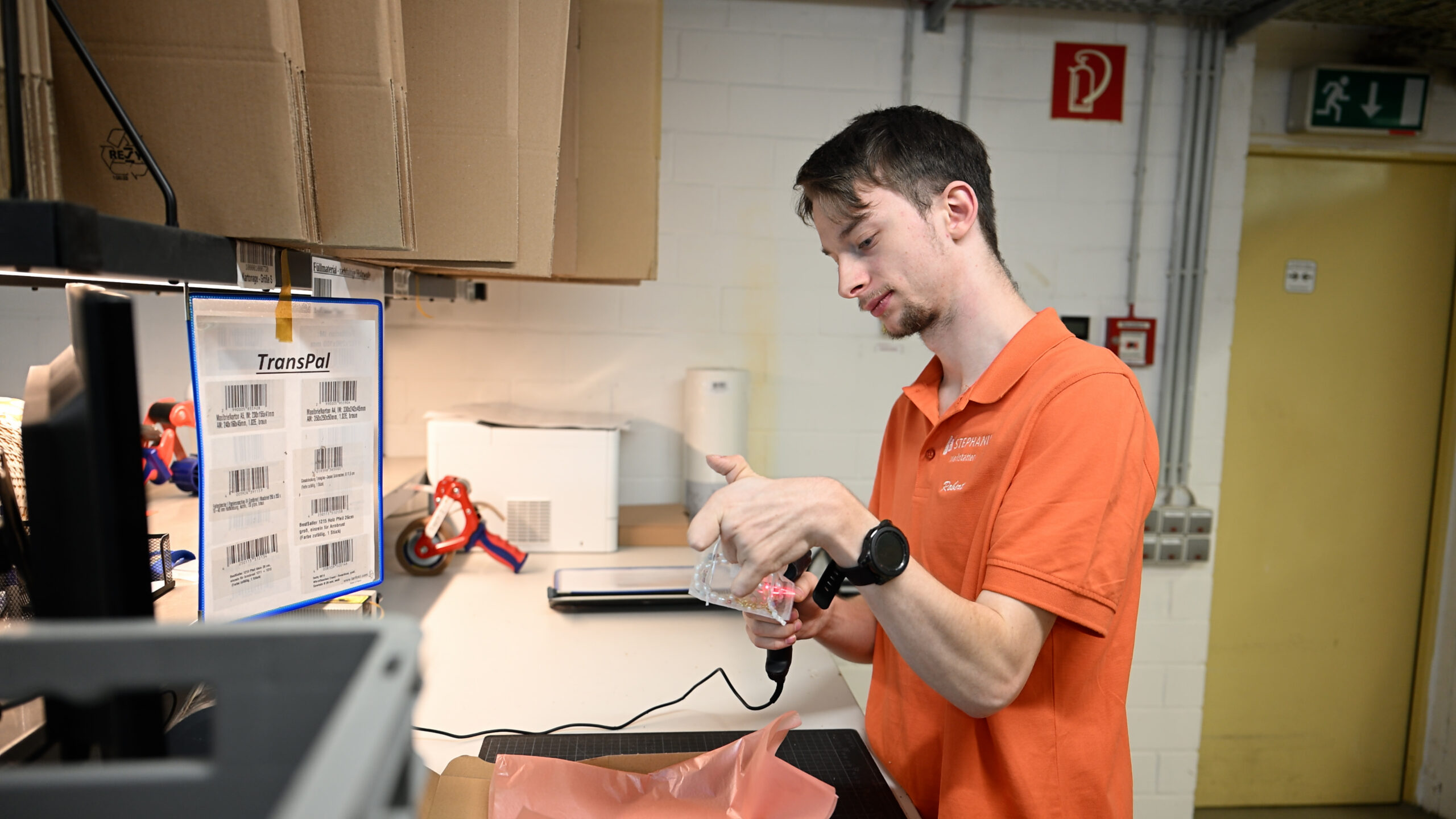 Ein Beschäftigter mit Einschränkungen scannt am Packtisch der Stephanus-Werkstätten einen Artikel. Er nutzt dabei die JTL-Software und trägt ein orangenes Poloshirt.