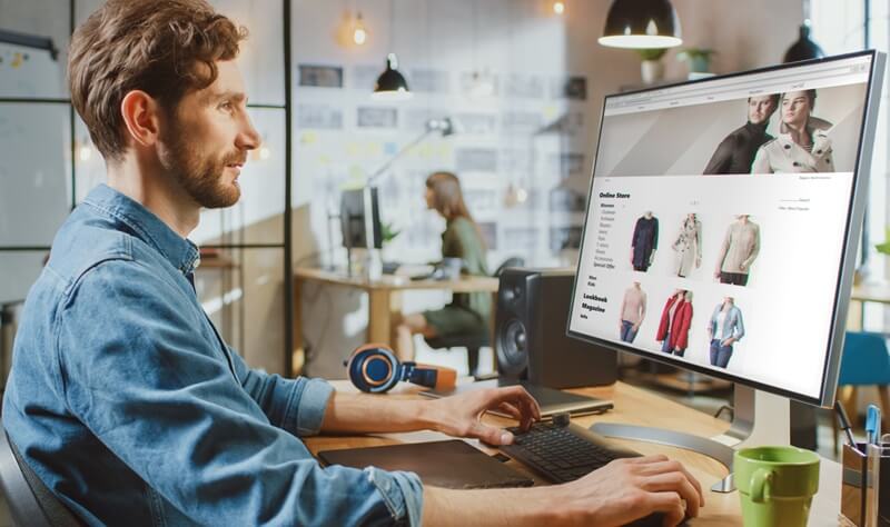 Mann im Jeanshemd am Schreibtisch im Großraumbüro betrachtet auf einem Monitor die Modeartikel eines Onlineshops, für die er mithilfe von KI-Tools Produktbeschreibungen erstellt.