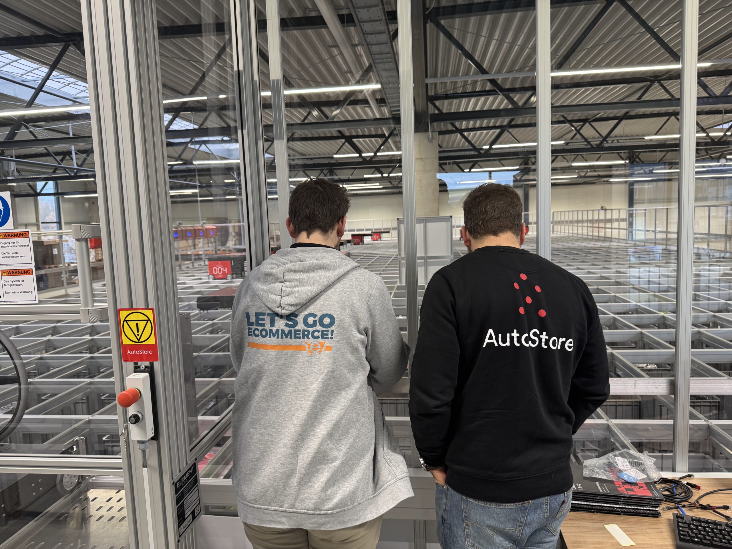 Zwei Maenner mit Pullovern von „AutoStore“ und „LET'S GO ECOMMERCE!“ blicken auf das automatisierte Lager. Warnschilder mit „AutoStore“ und Sicherheitsanweisungen sind an den Gittern angebracht.