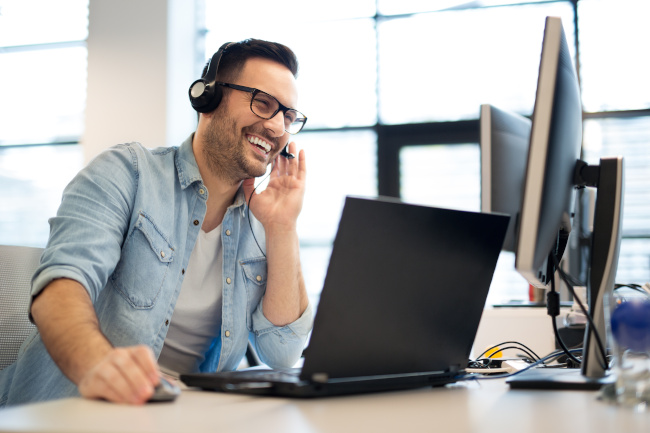 Junger Mann lacht am Schreibtisch mit Laptop und Headset