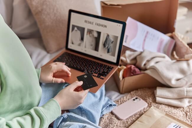 Eine Person sitzt auf einem Bett, hält eine schwarze Kreditkarte in der Hand und schaut auf einen Laptop, auf dem eine Website mit der Überschrift „RESALE FASHION“ und Bildern von Kleidung angezeigt wird. Daneben liegt ein geöffnetes Paket mit Kleidung und Versandunterlagen. Auf dem Bett liegen außerdem ein Smartphone und weitere Versandmaterialien.