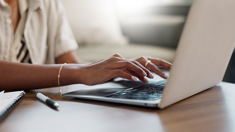 Nahaufnahme von Händen, die auf einer Laptop-Tastatur tippen. Auf dem Tisch liegen ein Notizbuch und ein Stift. Die Umgebung wirkt hell und ruhig, im Hintergrund ist ein unscharfes Sofa zu erkennen.