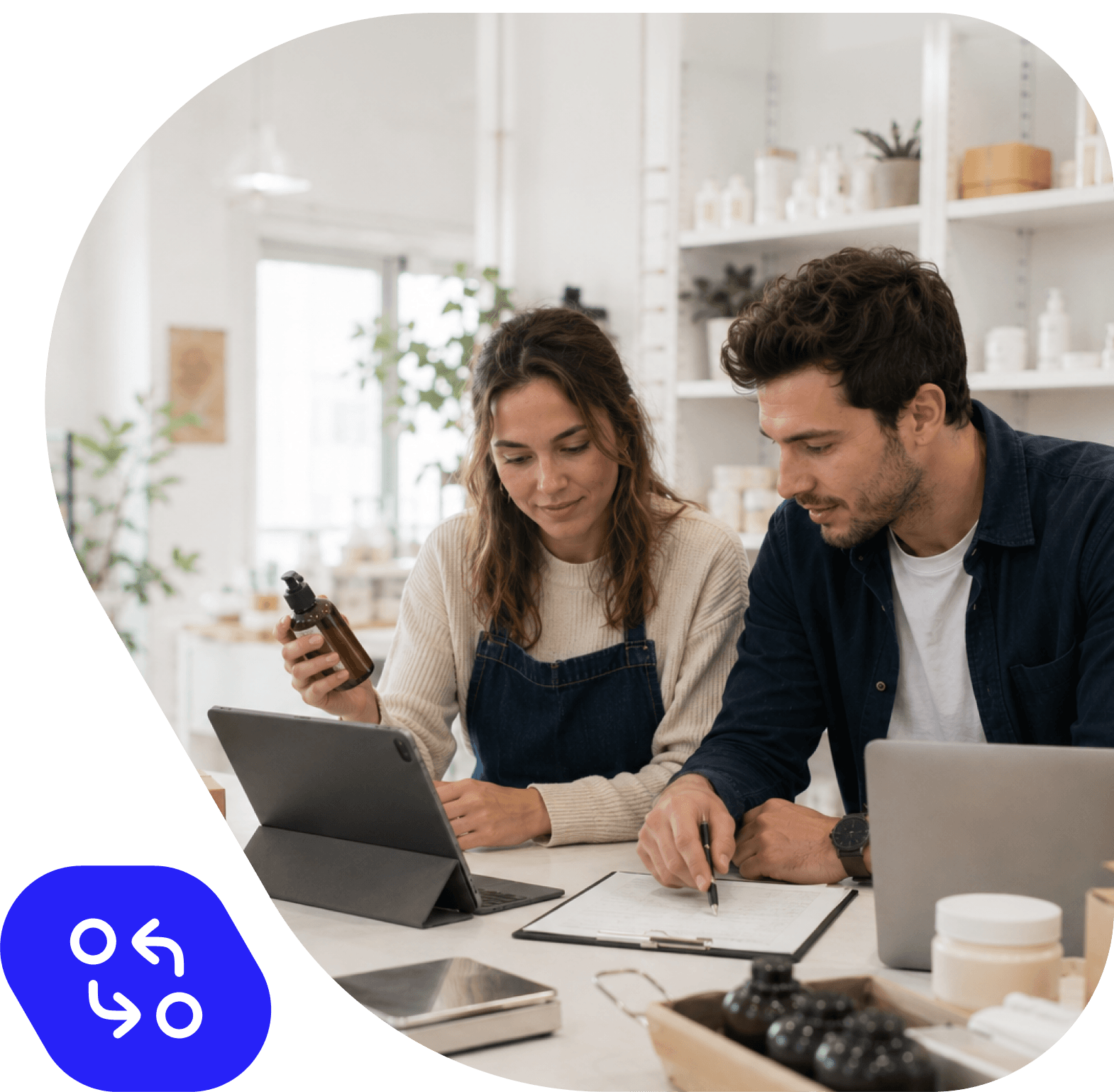 Zwei Personen arbeiten gemeinsam an einem Tisch mit Tablet, Laptop und Unterlagen in einem hellen Kosmetik- oder Naturkostladen. Die Frau hält ein braunes Fläschchen in der Hand. Unten links ein blaues Icon mit Pfeilen, das Datenaustausch symbolisiert.